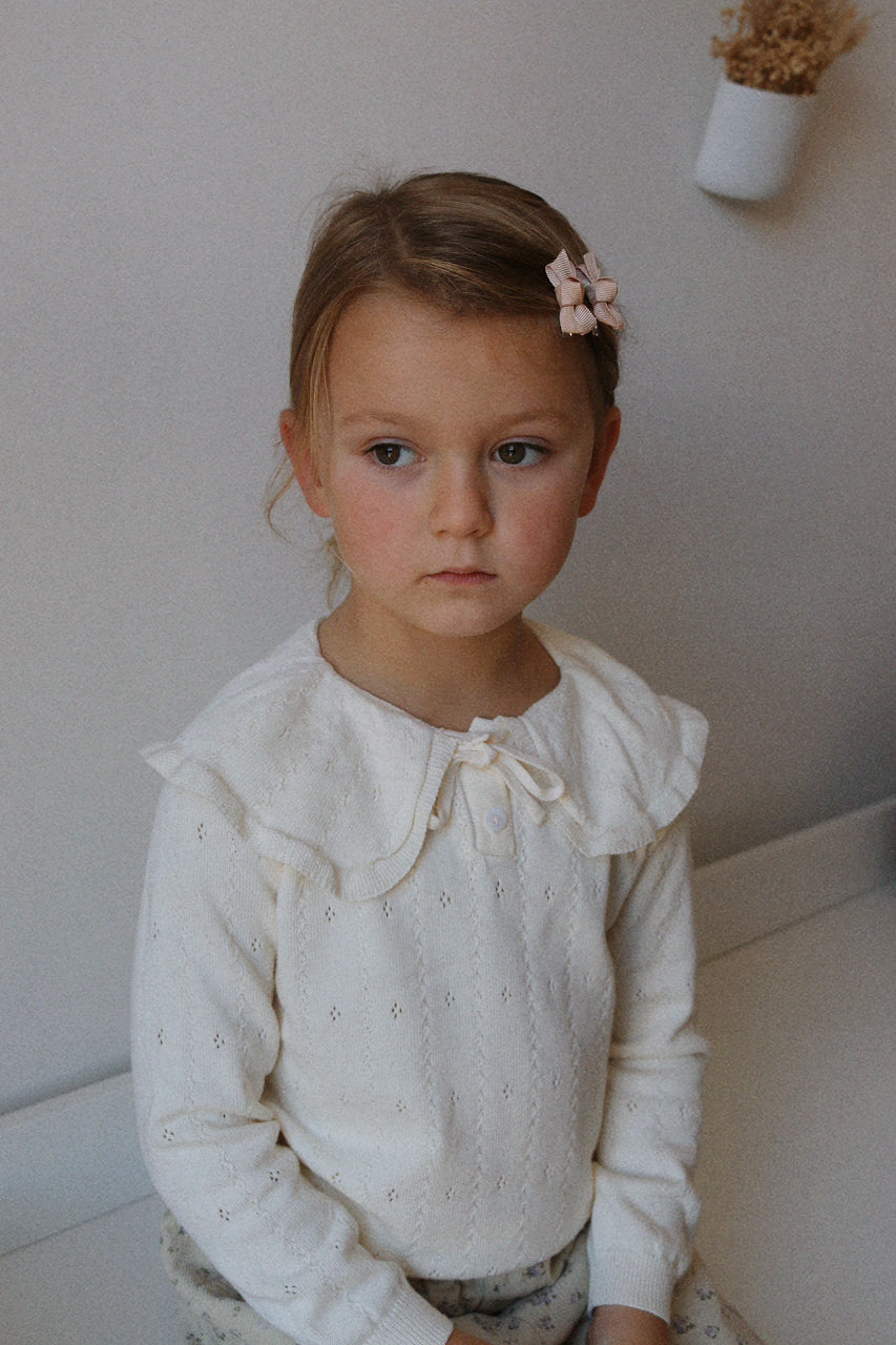Young girl wearing a ivory jumper with ruffled details, sitting against a neutral background.