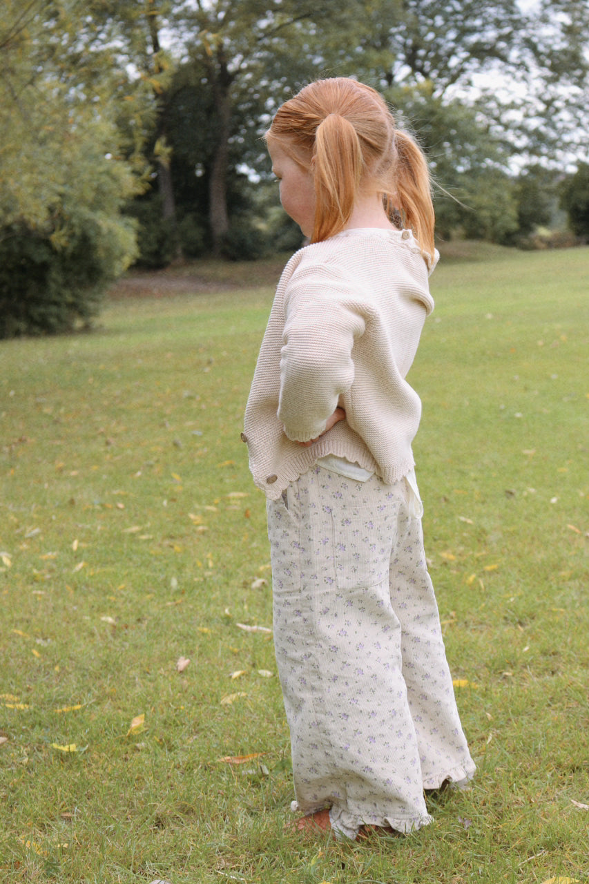 Person wearing a light-colored cardigan and floral trousers standing in a grassy field with trees in the background.