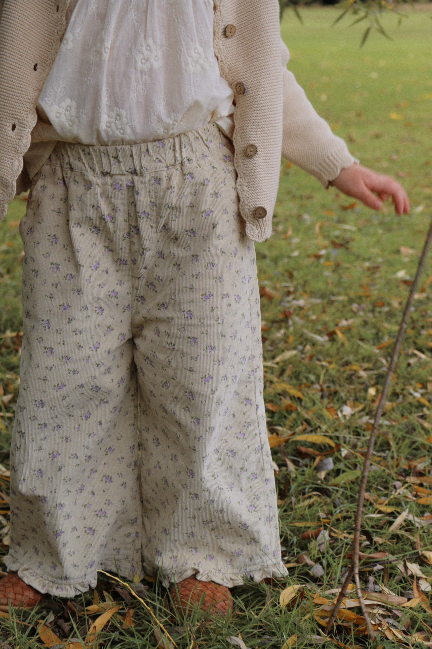 Girl wearing a beige cardigan and floral pants standing in a grassy area with fallen leaves.