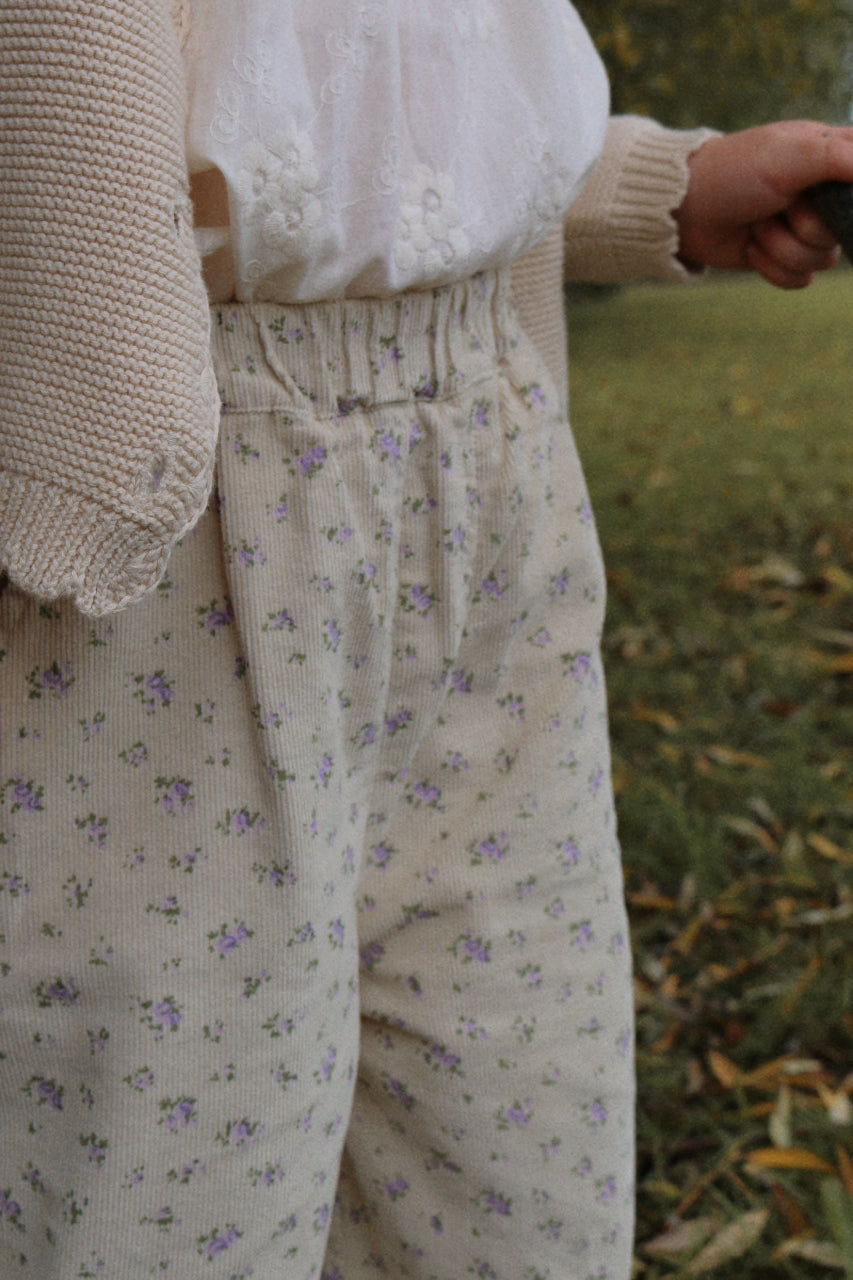 Girl wearing a beige cardigan and floral corduroy pants standing outdoors on grass.