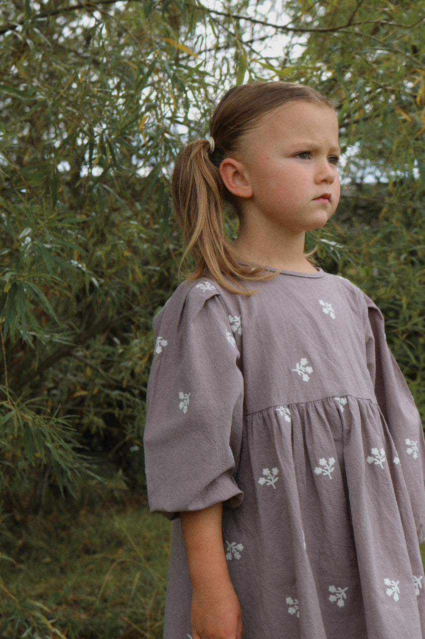 Young girl in a gray dress with floral patterns standing outdoors against a green background