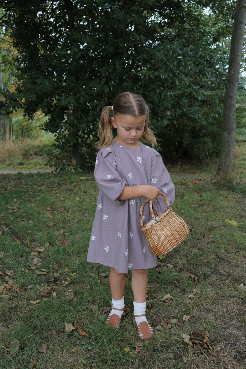 Young girl in a purple dress holding a woven bag in a natural setting