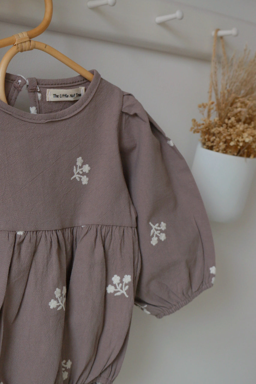 grey children's romper with floral embroidery on a hanger against a white wall.