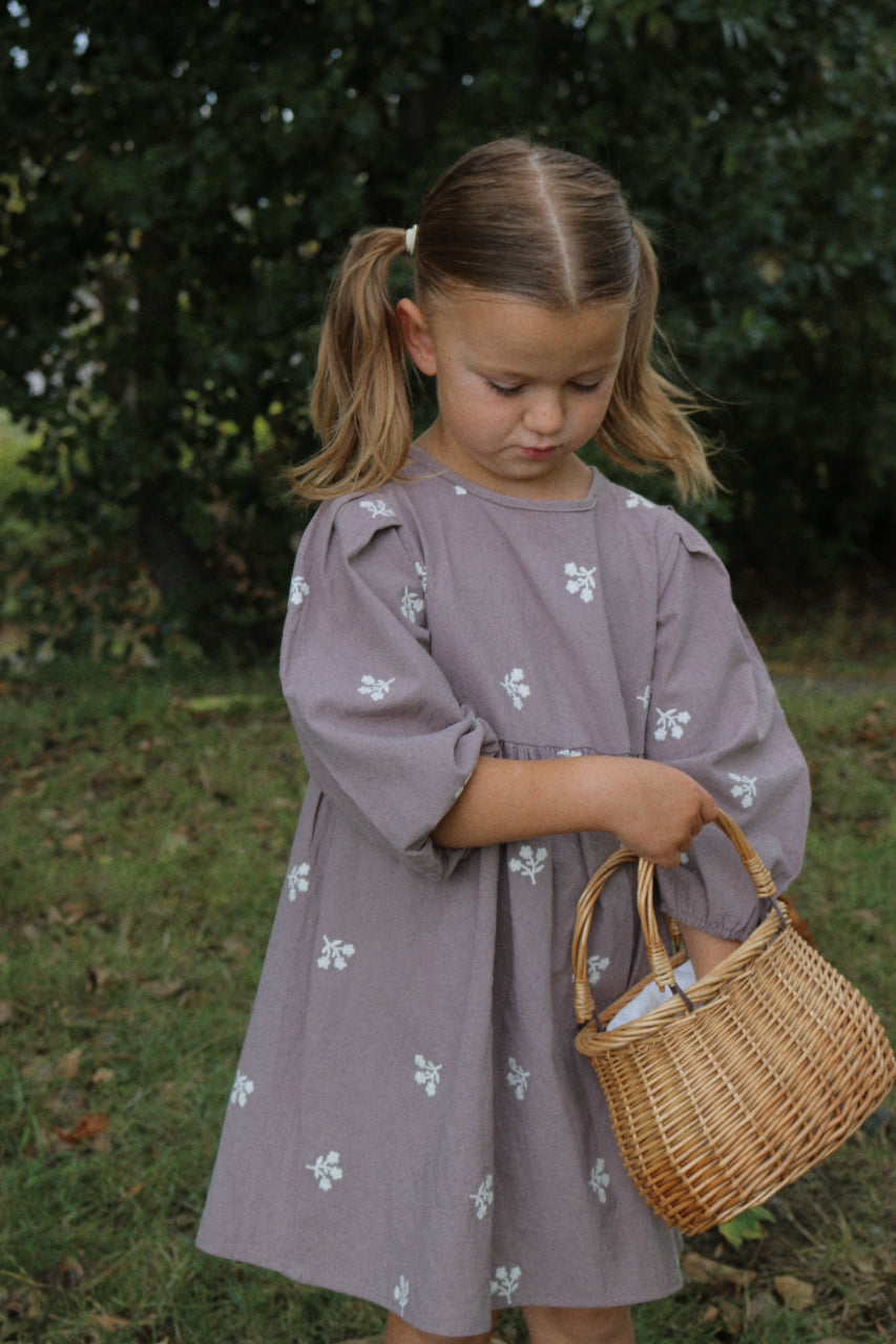 Young girl in a purple dress holding a wicker basket outdoors.