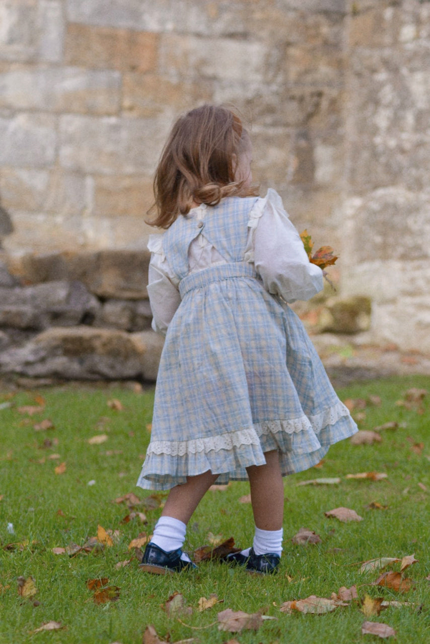 Child in a checkered dress standing on grass with a stone wall in the background