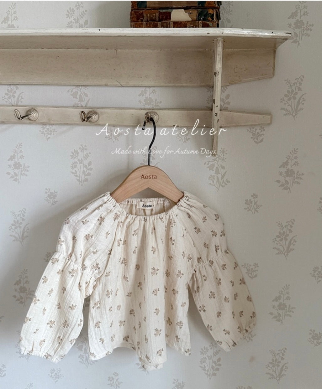 Floral blouse hanging on a wooden hanger against a floral wallpapered wall.