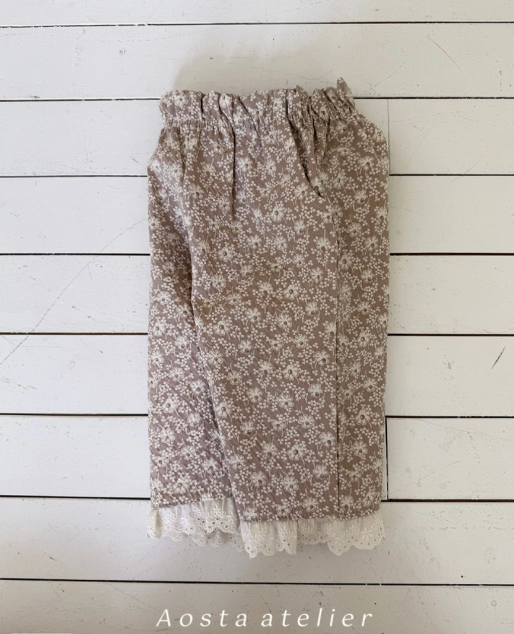 Floral patterned pants with lace trim on a white wooden background, branded 'Aosta atelier'.