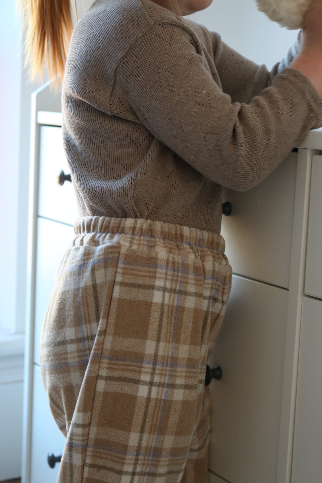 Person wearing a brown sweater and plaid pants standing in front of white cabinets.