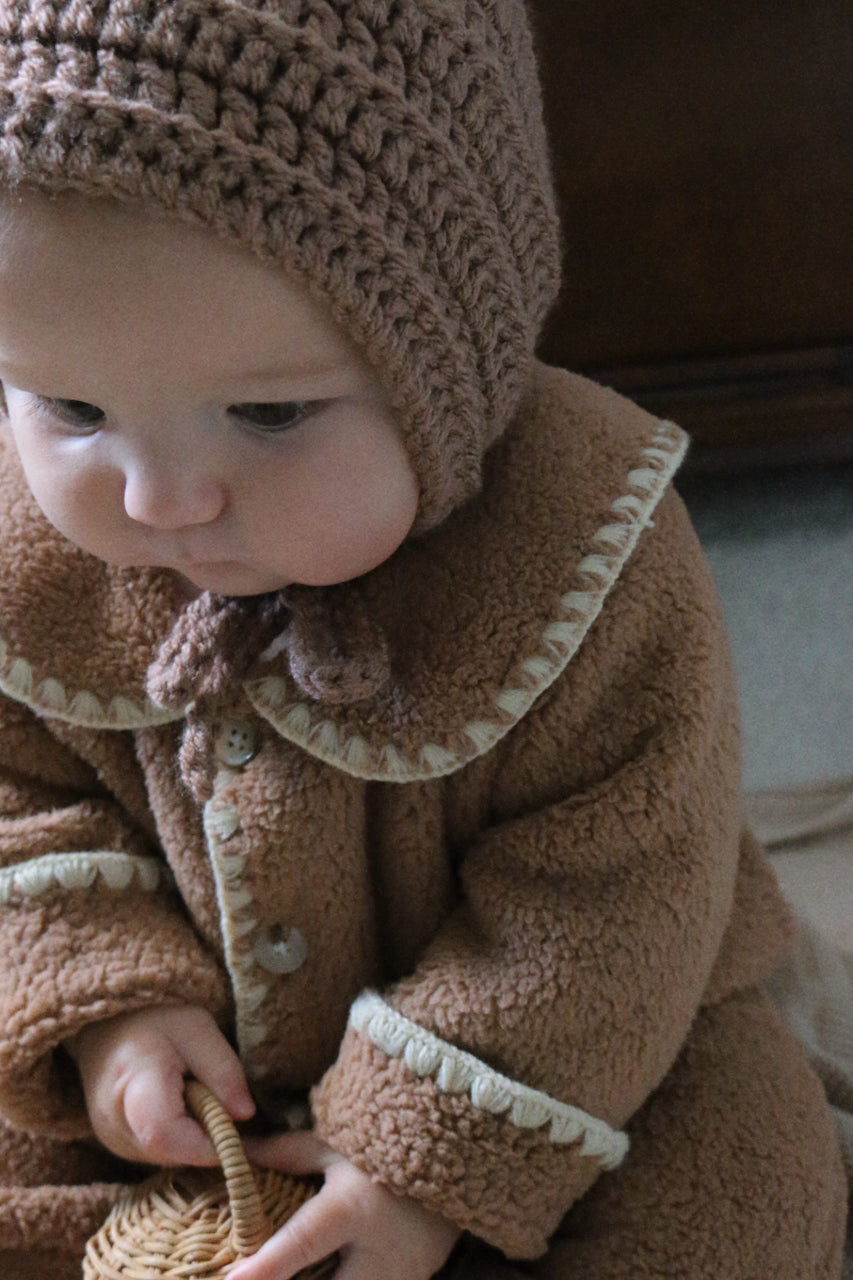 Baby wearing a brown coat and knitted hat holding a small woven basket.