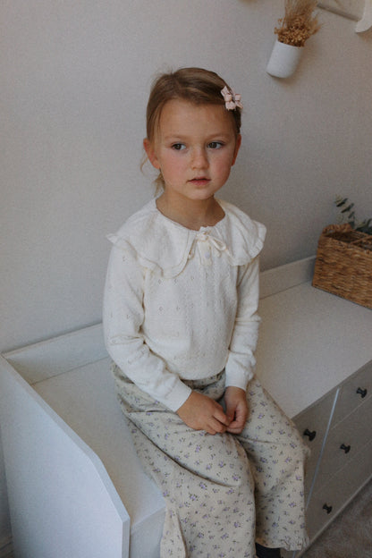 Young girl in a cream jumper and beige corduroy pants with ruffled details sitting on white drawers 