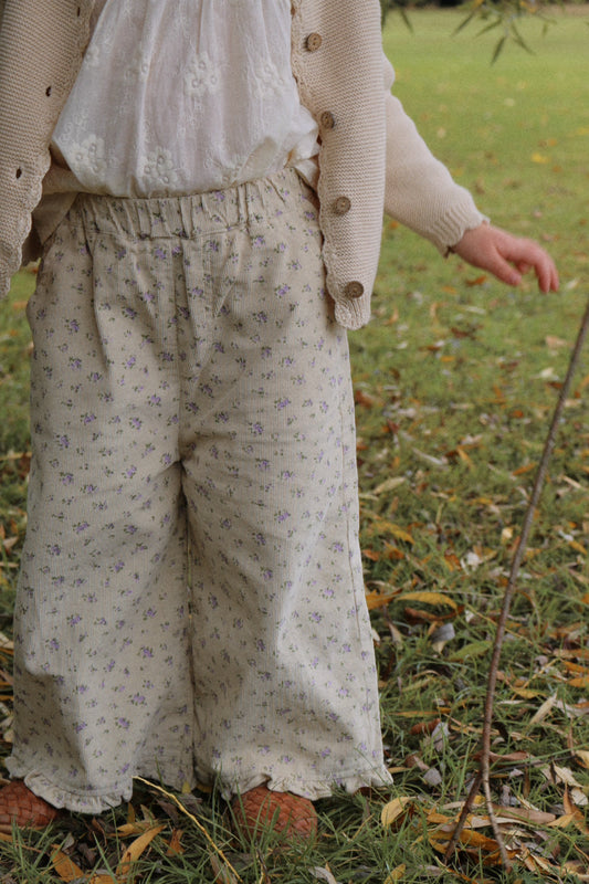 Girl wearing a beige cardigan and floral pants standing in a grassy area with fallen leaves.