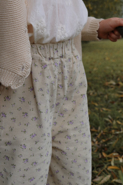 Girl wearing a beige cardigan and floral corduroy pants standing outdoors on grass.