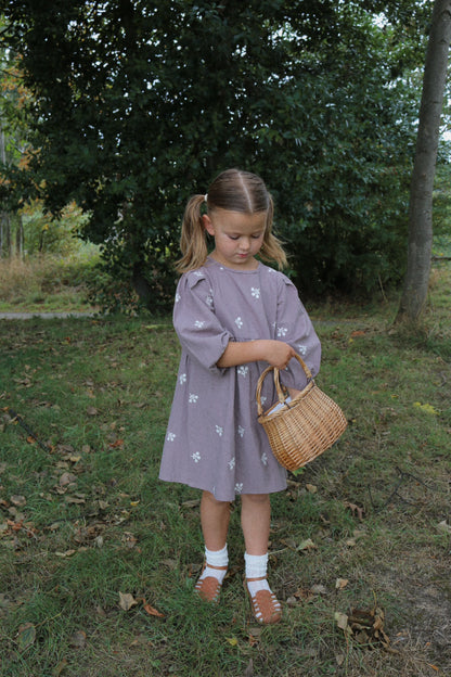 Young girl in a purple dress holding a woven bag in a natural setting