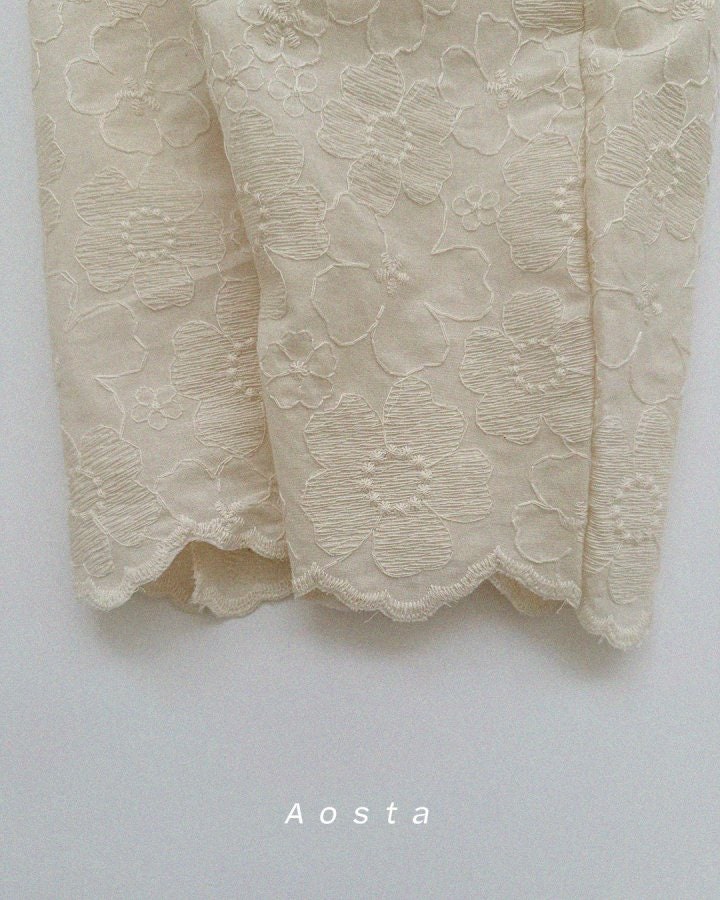 Close-up of beige lace pant cuffs with 'Aosta' branding on a light gray background