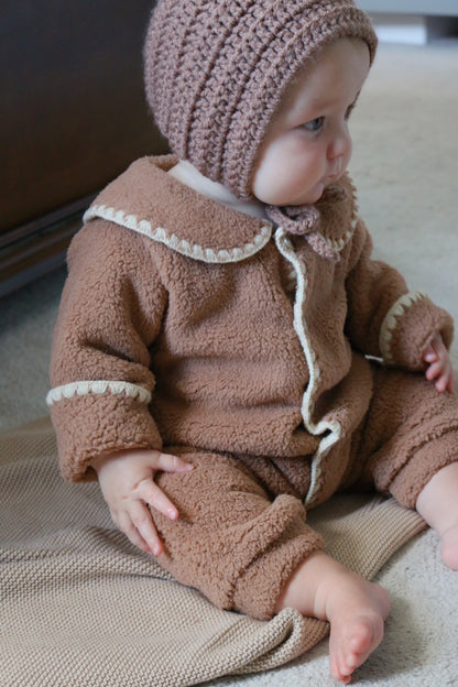 Baby in a brown outfit with a matching bonnet sitting on a blanket.