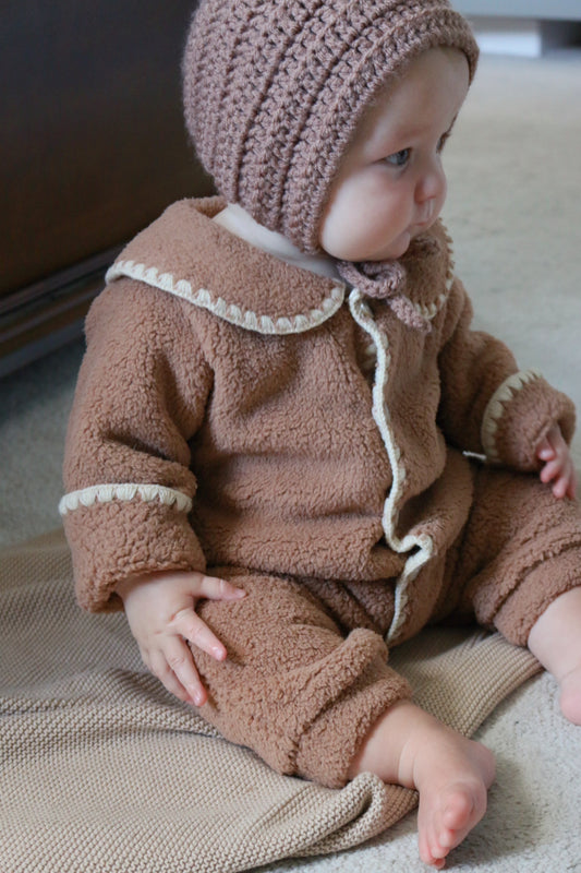 Baby in a brown outfit with a matching bonnet sitting on a blanket.