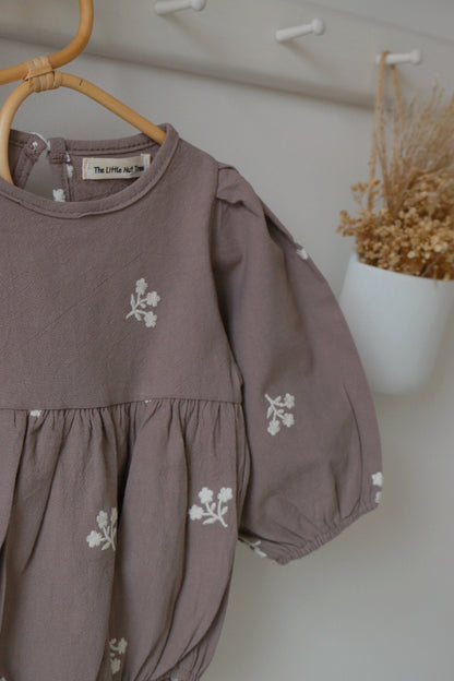 grey children's romper with floral embroidery on a hanger against a white wall.
