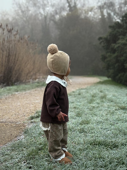 Child wearing a brown coat and beige hat with a pom-pom, standing on a frosty path.