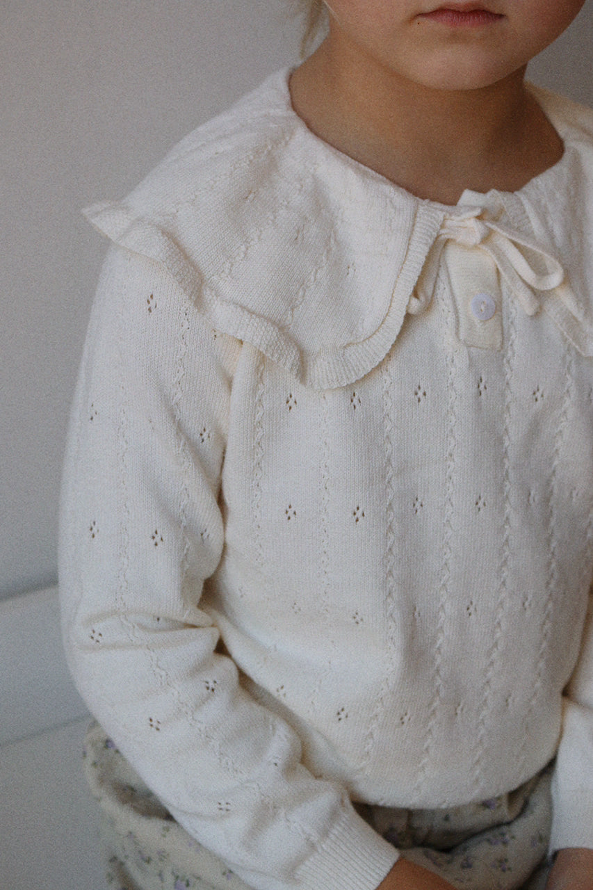 Young girl wearing a ivory jumper with ruffled details, sitting against a neutral background.