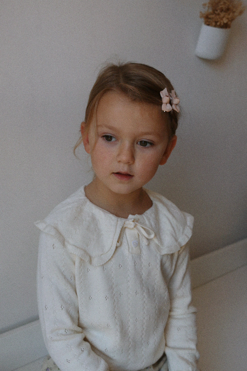Young girl wearing a ivory pointelle jumper with ruffled collar against a plain wall.