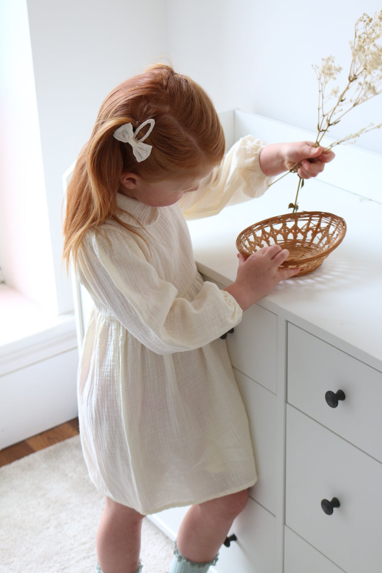 Child holding a small woven basket with dried flowers indoors wearing cream dress