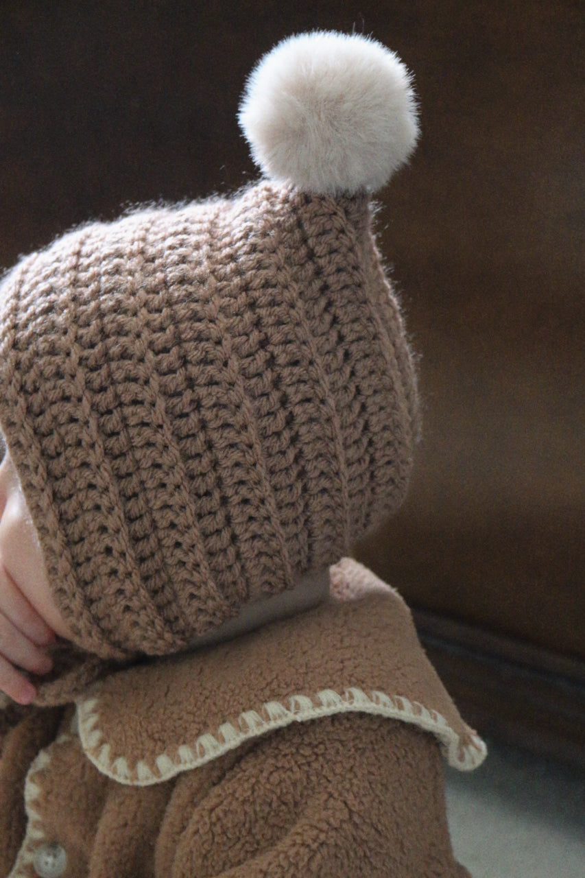 Brown knitted crochet pixie bonnet worn by baby with a beige pom-pom on a blurred background
