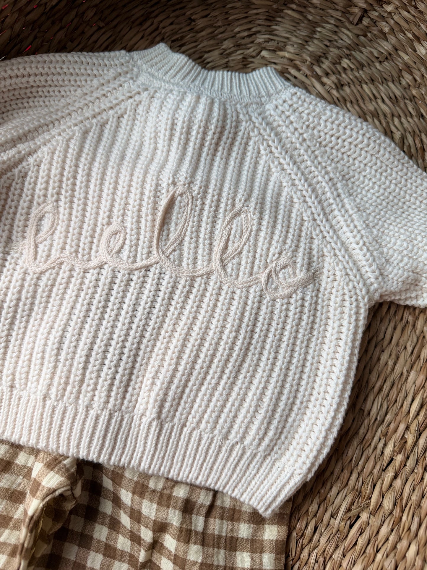 Ivory knitted cardigan, with the wording 'Hello' embroidered on the back. Flat laid on a wicker basket. 