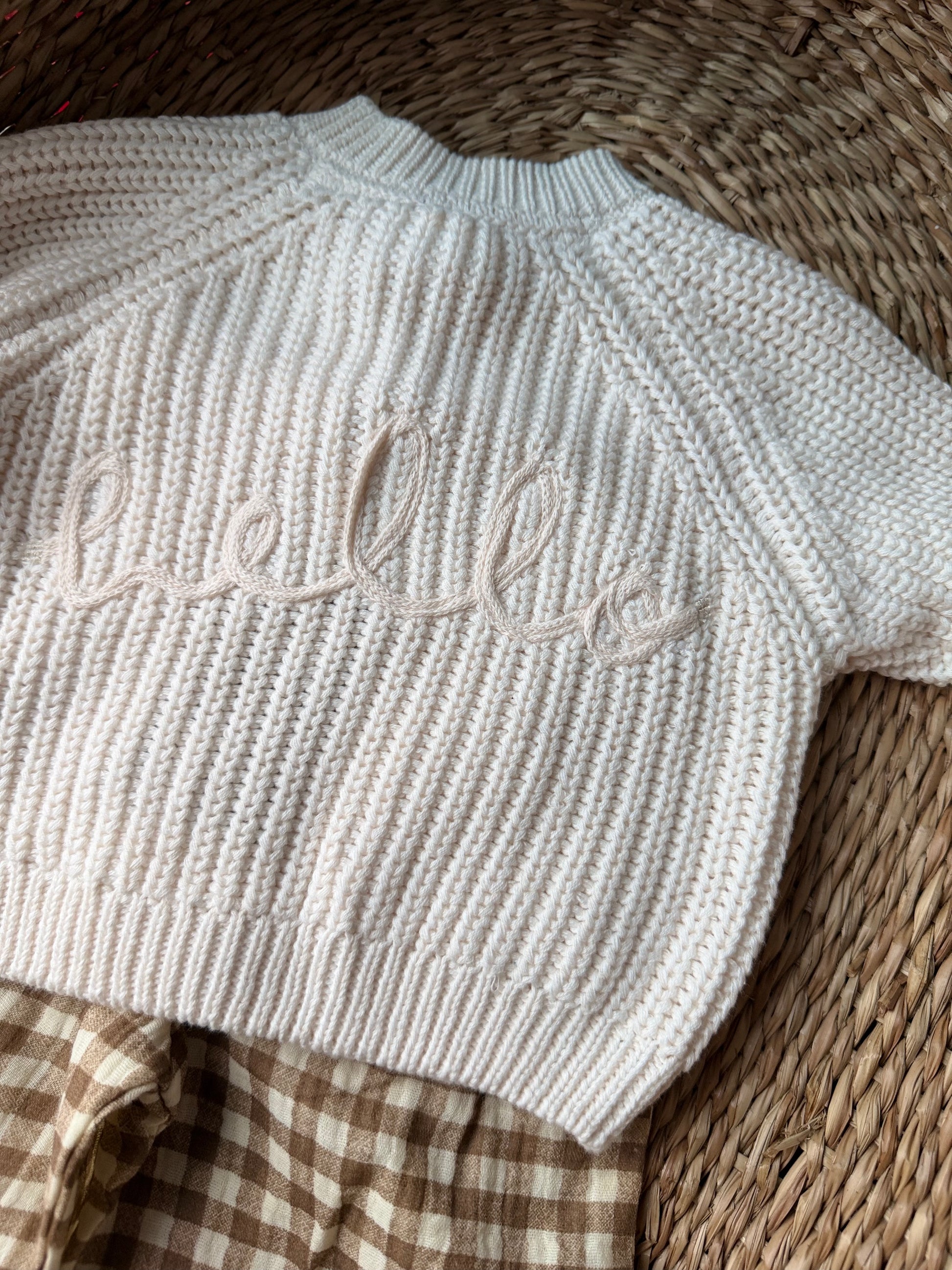 Ivory knitted cardigan, with the wording 'Hello' embroidered on the back. Flat laid on a wicker basket. 