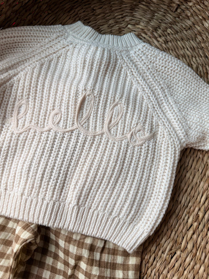 Ivory knitted cardigan, with the wording 'Hello' embroidered on the back. Flat laid on a wicker basket. 