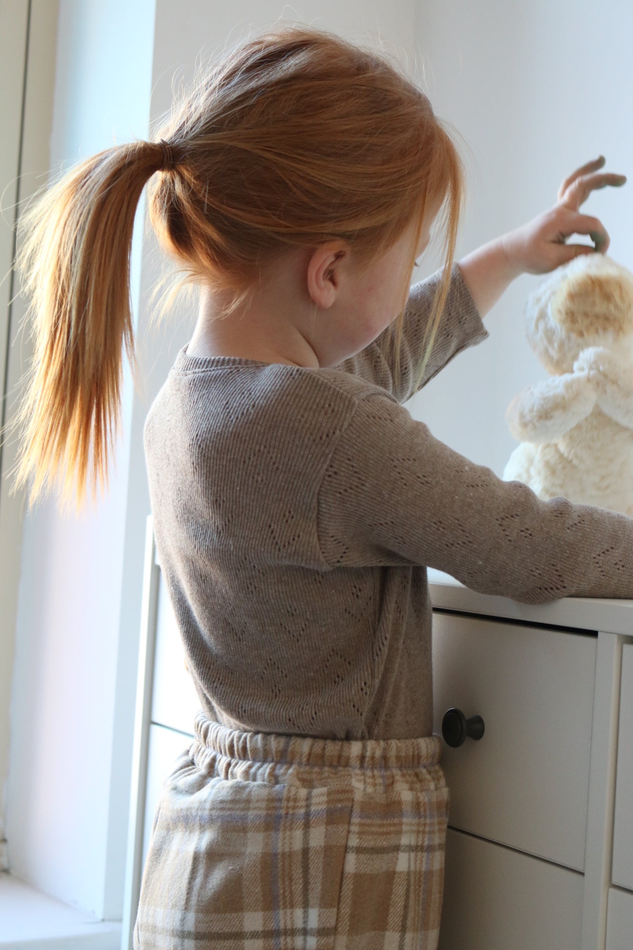 Child playing with a teddy bear in a room with white walls and a window.