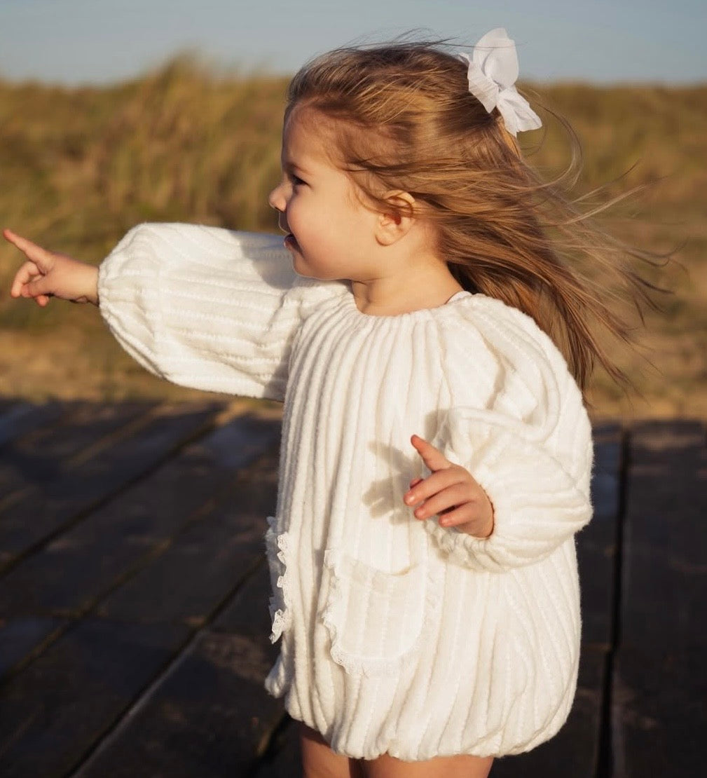 Child in a white outfit standing on a wooden boardwalk with grassy dunes in the background