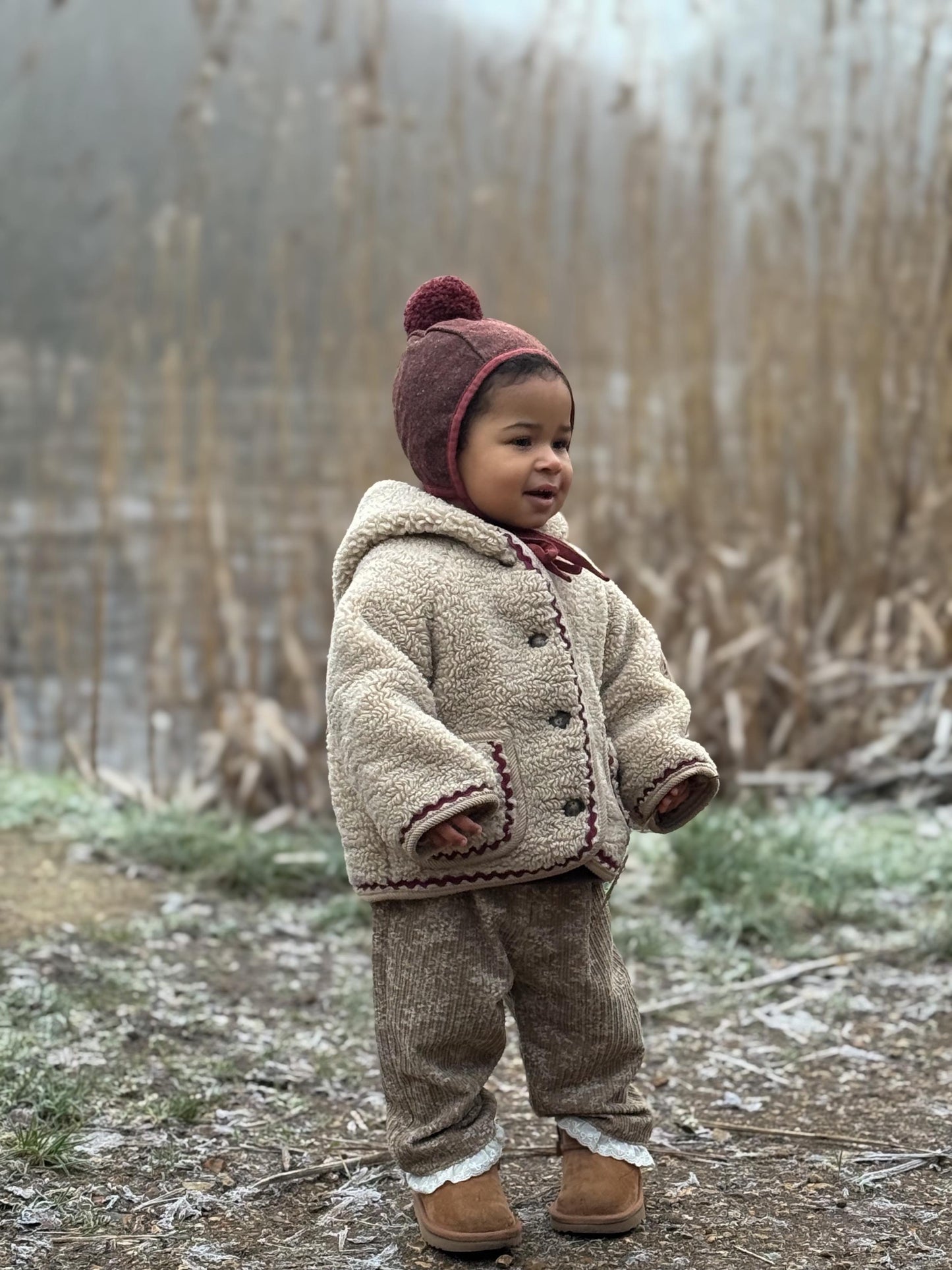 Child wearing a beige coat and brown pants standing in a natural setting with tall grass.