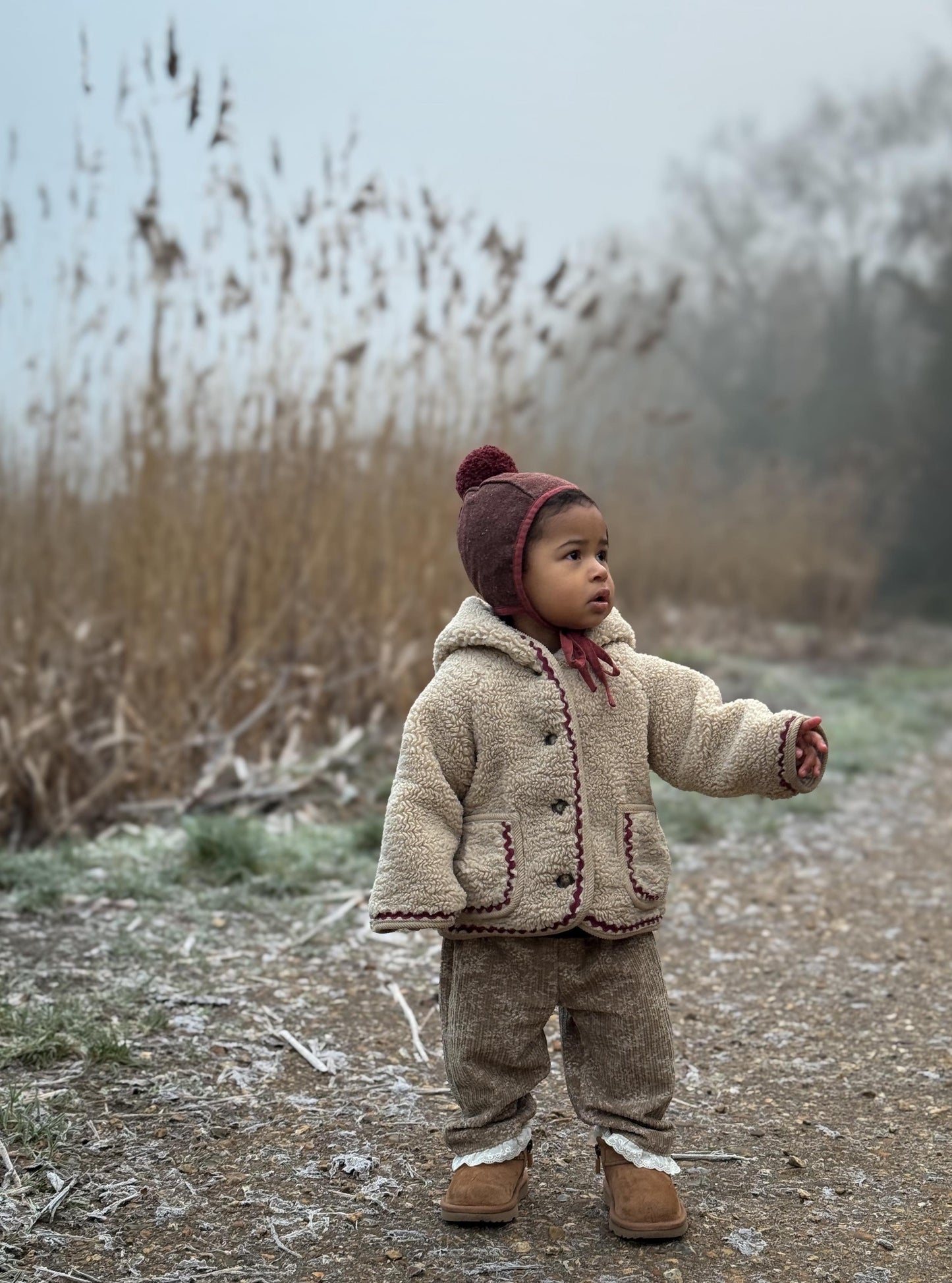 Child in a beige coat and brown hat standing in a frosty field with tall grass and bare trees.