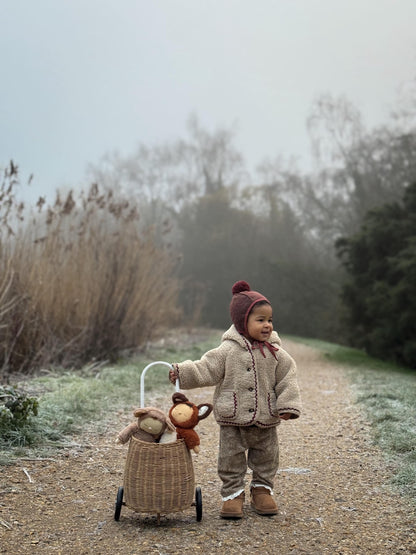 Child in a coat and hat pushing a toy stroller with a teddy bear in a natural setting.