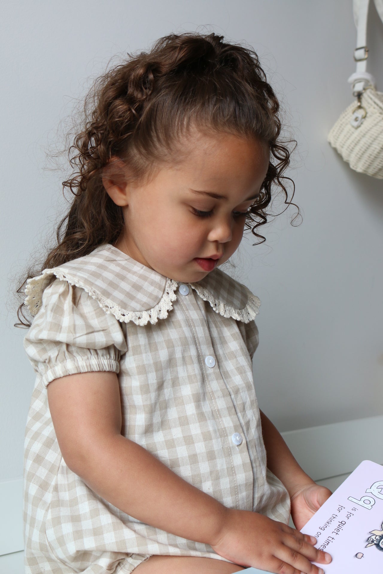 Child in a checkered romper holding a book on a light-colored drawers