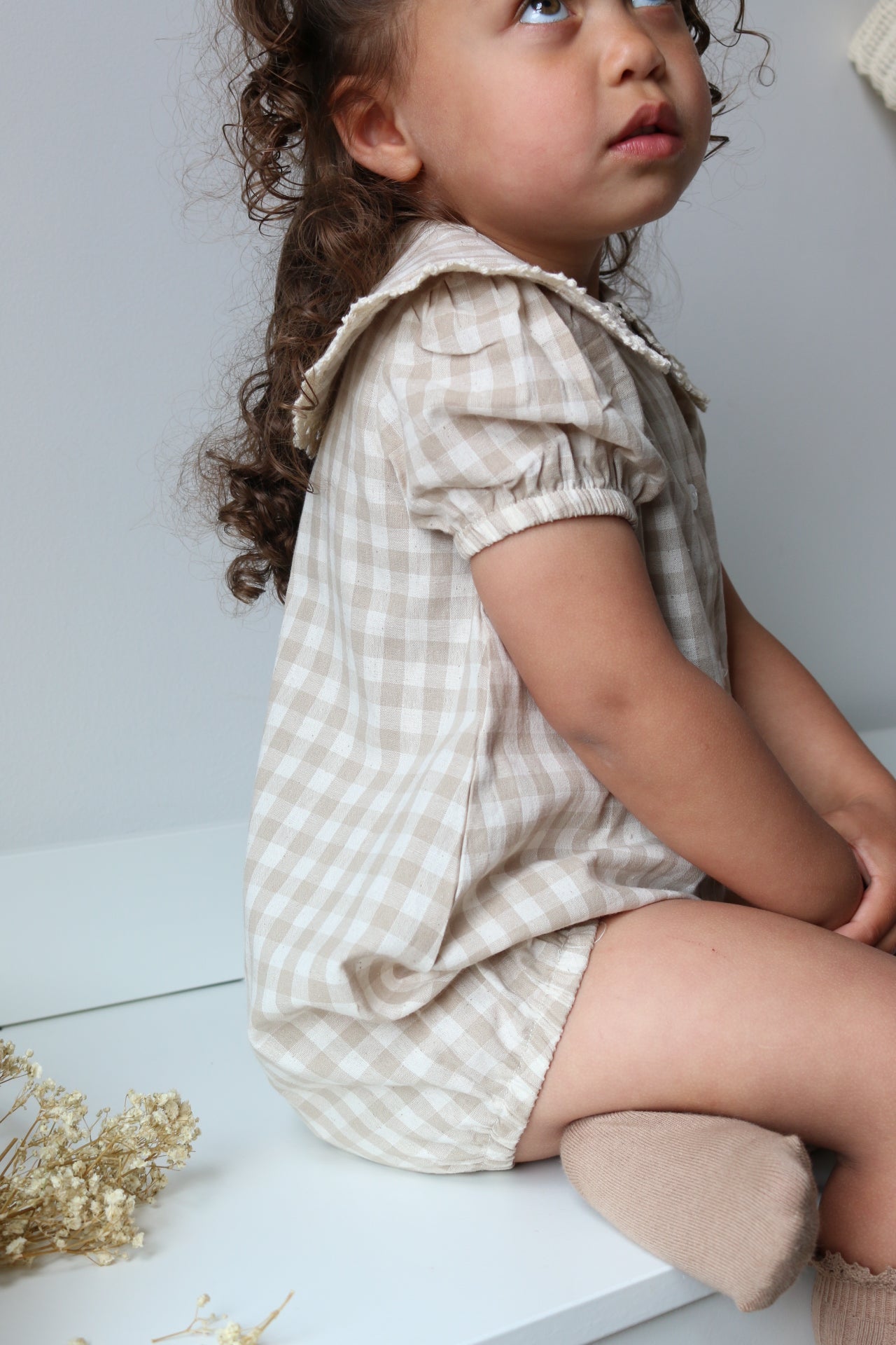Child wearing a checkered romper lying on a light surface with dried flowers.