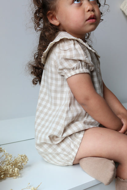 Child wearing a checkered romper lying on a light surface with dried flowers.