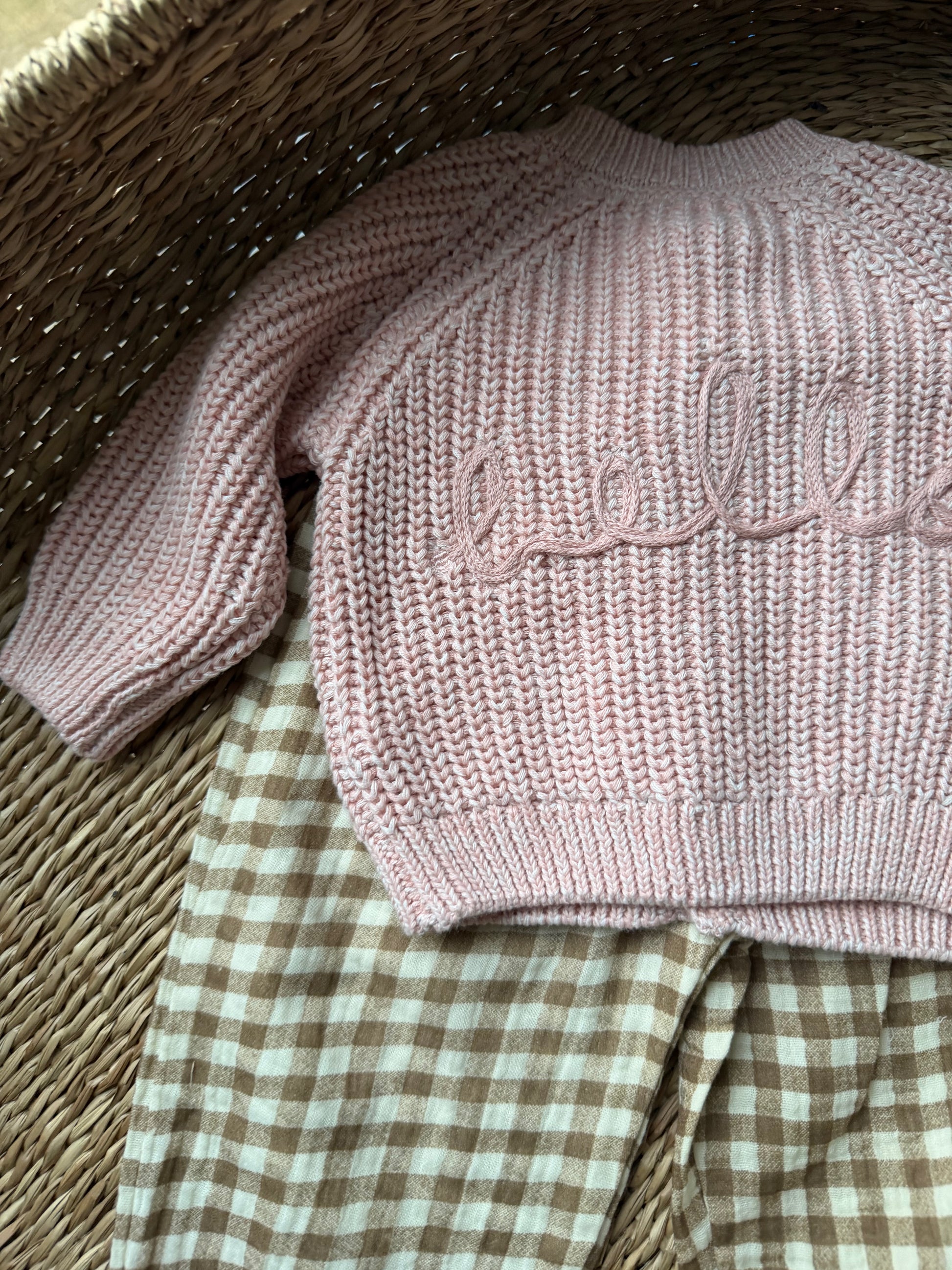 Pink knitted cardigan with 'hello' embroidery with checkered pants flat laid on a wicker basket.