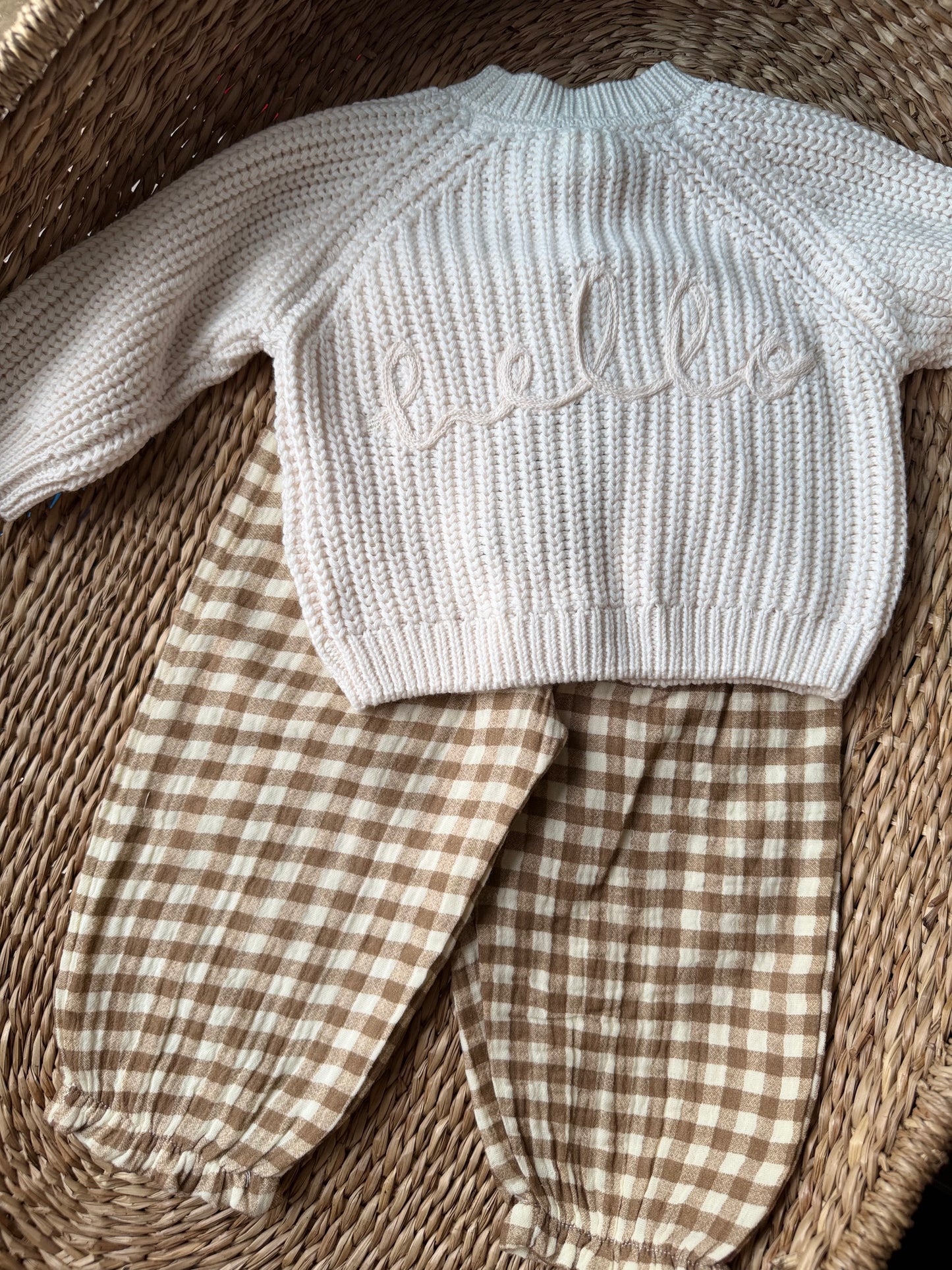 Knitted baby cardigan with checkered pants flat laid on a woven surface