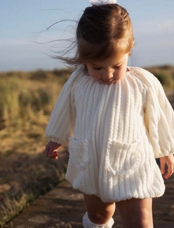 Child wearing a white velour romper standing on a beach.