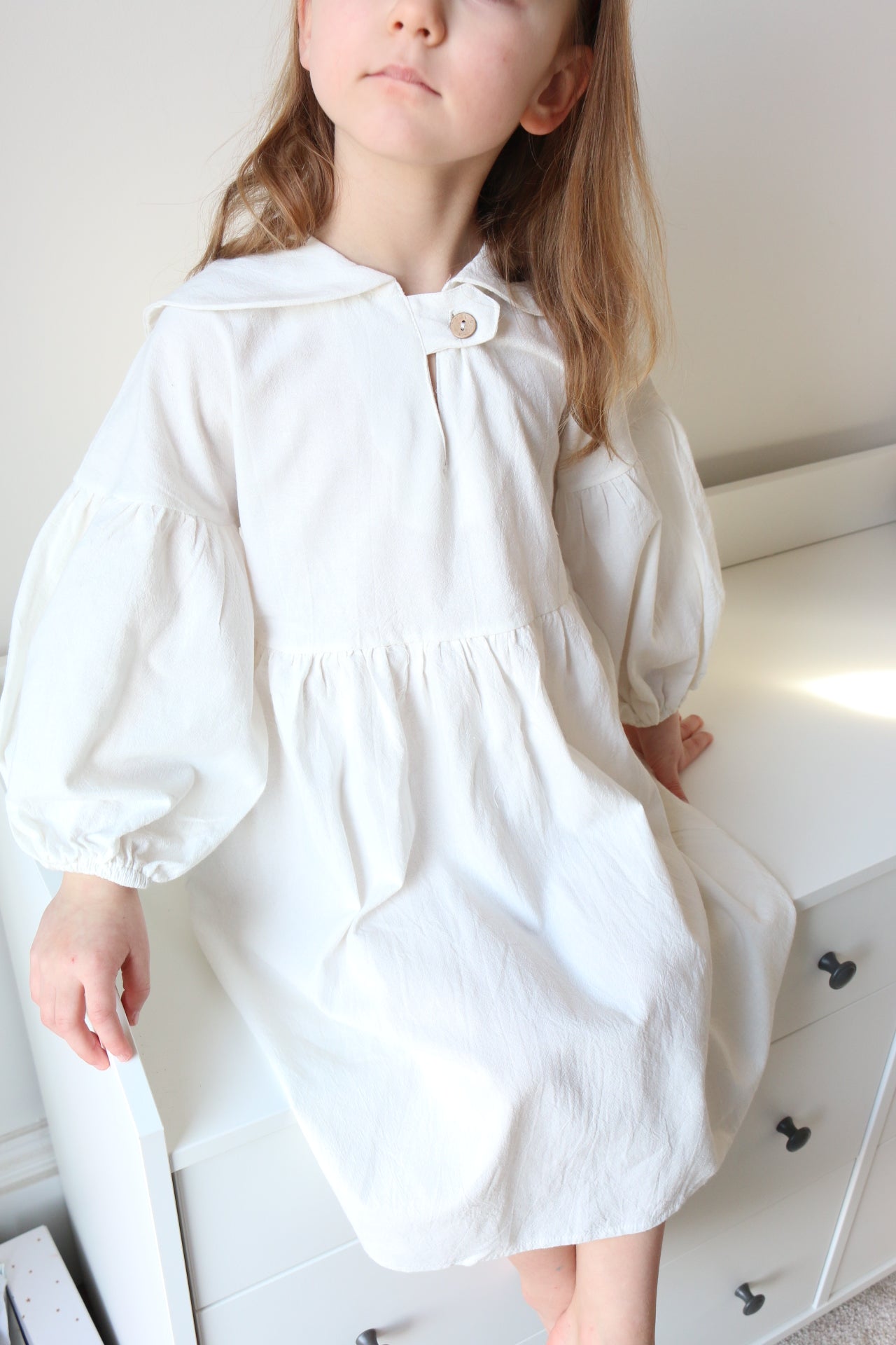 Girl sitting lying on a white drawers wearing a white dress with a button detail.