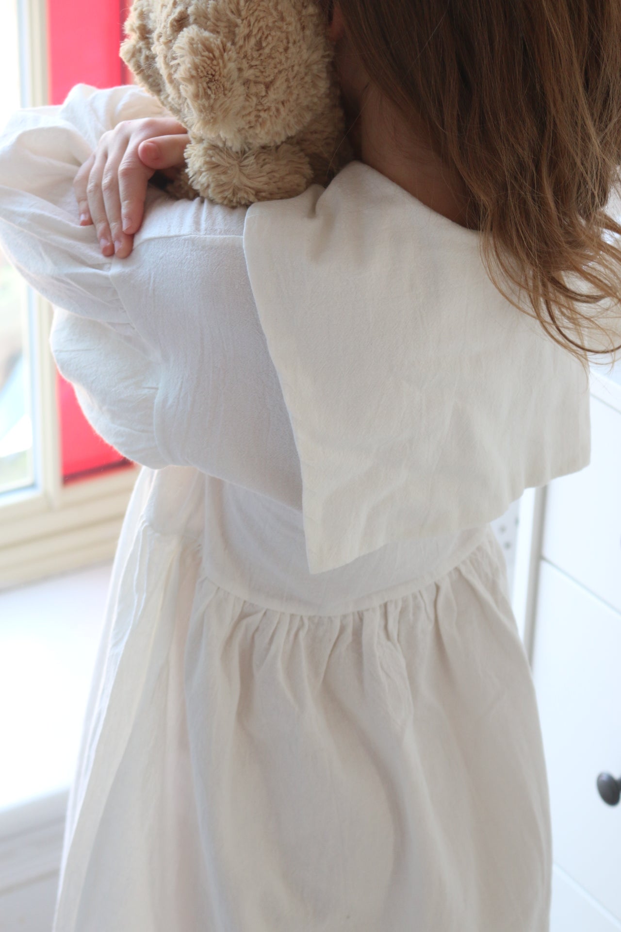 girl wearing a white dress holding a teddy bear indoors.