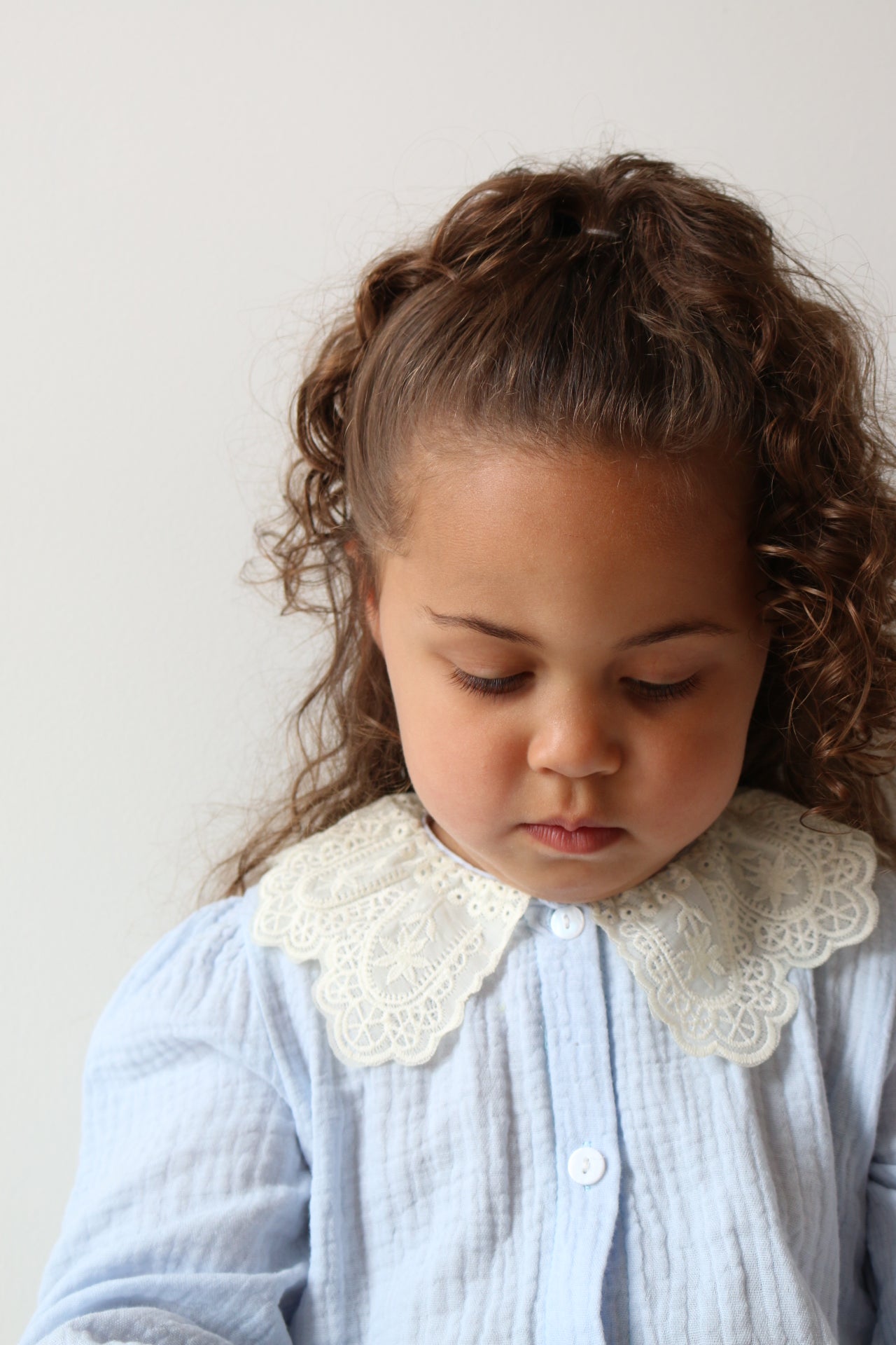 Child with curly hair wearing a light blue romper with lace details on a white background